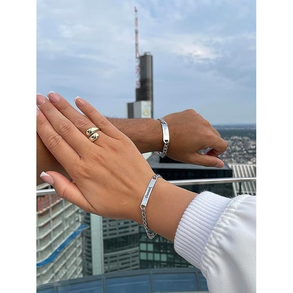 Männer- und Frauenhände mit personalisierten Partnerarmbändern, die Namen des Partners tragen, vor einer Skyline im Hintergrund.