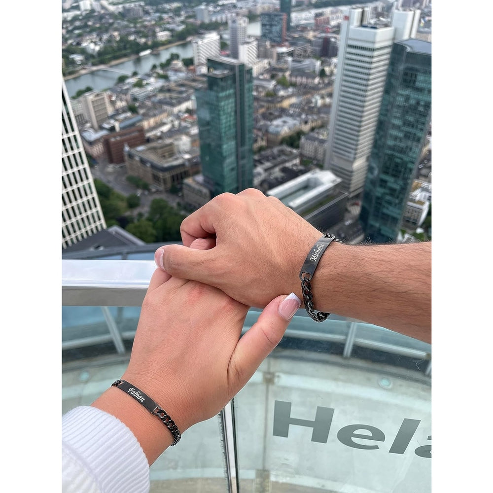 Paar mit Partnerarmbändern: Männer- und Frauenhand tragen Armbänder mit dem Namen des Partners, Skyline im Hintergrund.