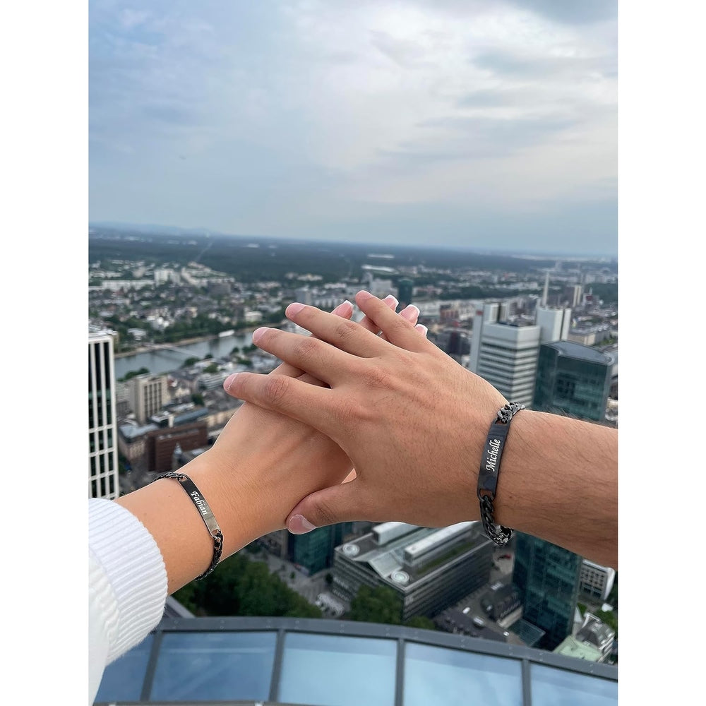 Hand in Hand: Männer- und Frauenhand mit personalisierten Partnerarmbändern, Skyline als Hintergrund.