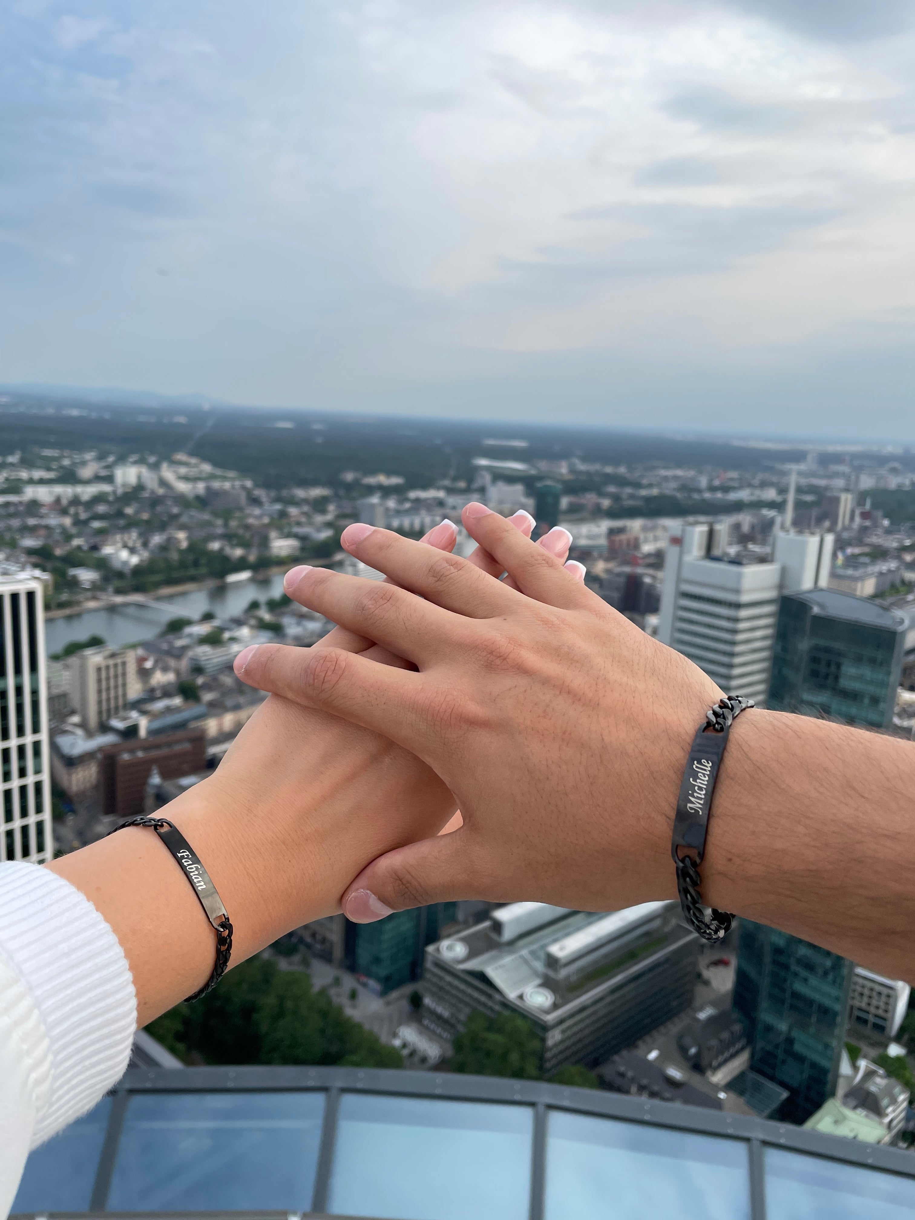 Männer- und Frauenhände mit personalisierten Partnerarmbändern, die Namen des Partners tragen, vor einer Skyline im Hintergrund.